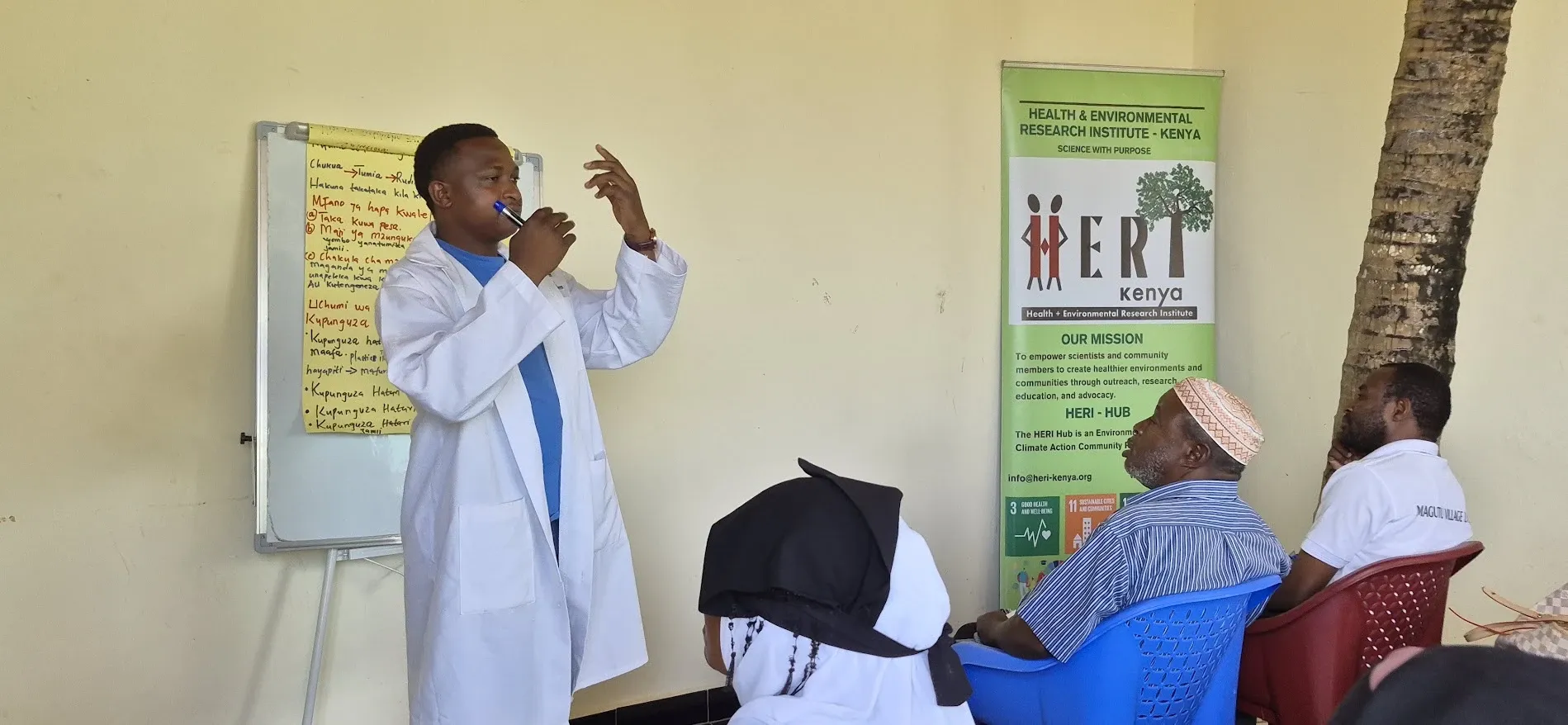 A HERI trainer in a lab coat leads a community training session in Kiswahili beside the HERI-Kenya banner